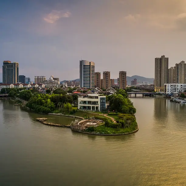 A small green island with a modern white building is surrounded by a river, with a city skyline and hills in the background under a partly cloudy sky at sunset.