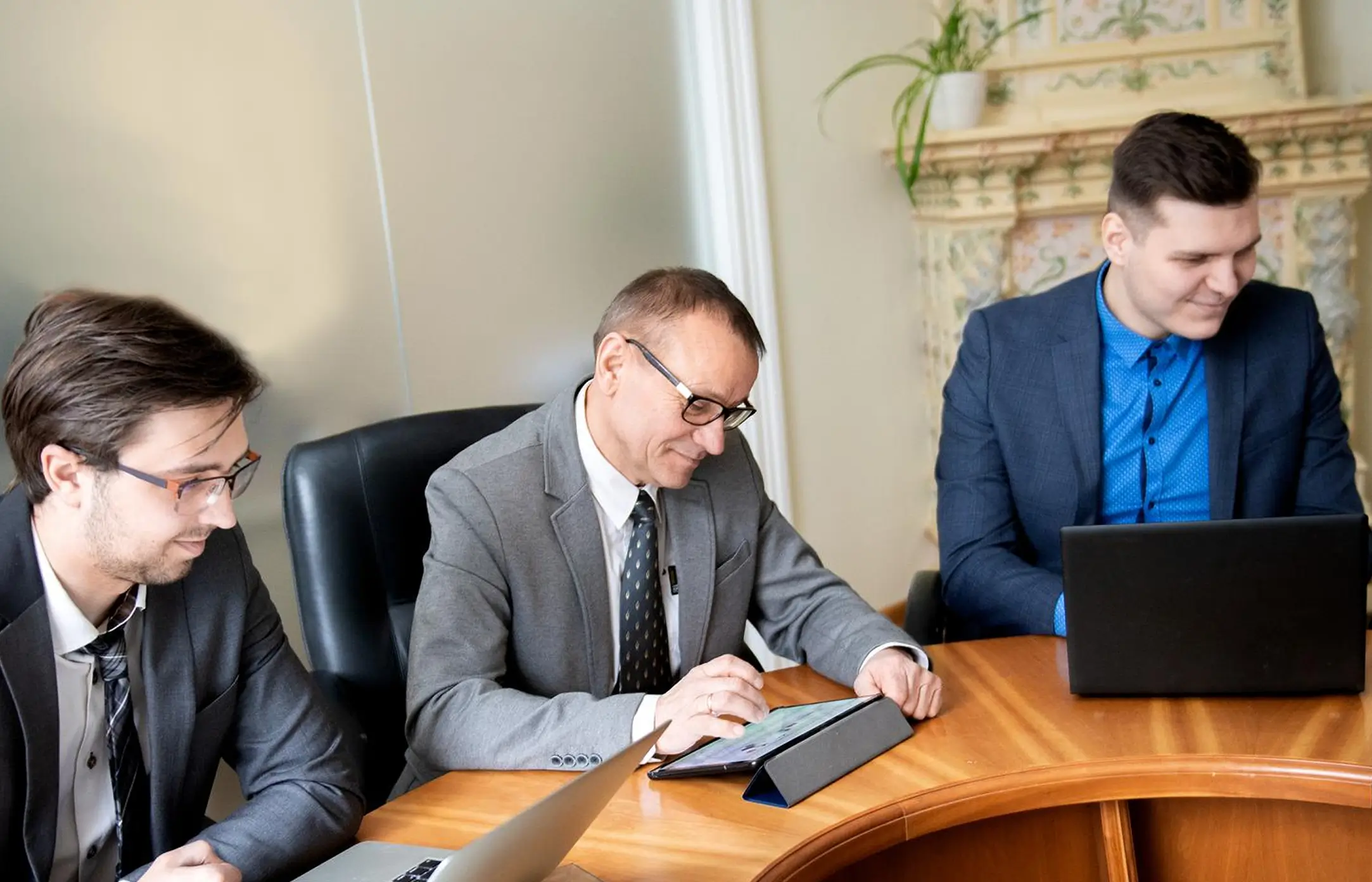 Three men in business attire working at a wooden table, with one using a tablet, another a laptop, and the third smiling while looking at his device in an office setting.
