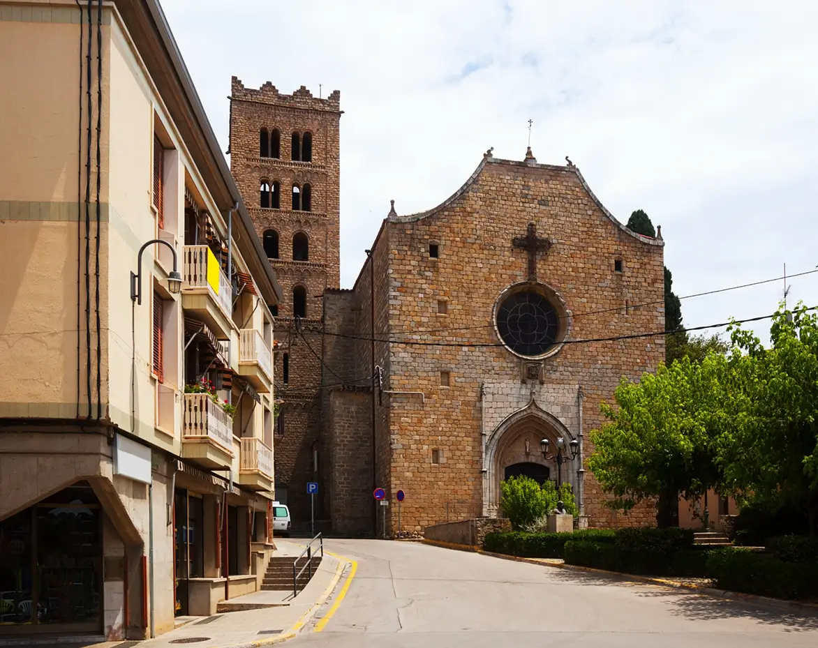 Street view of old stone church with large circular stained-glass window and cross above the entrance, adjacent to a tall rectangular bell tower with multiple arched openings, next to residential buildings with balconies and shuttered windows, under partly cloudy sky.