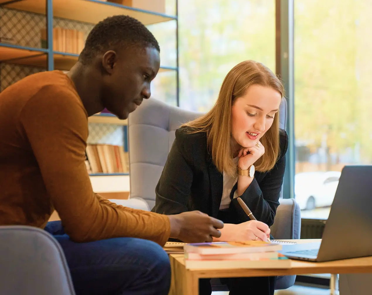 A man in a brown long-sleeve shirt and a woman in a black blazer sitting at a table with a laptop, books, and notebooks, engaged in discussion or study.