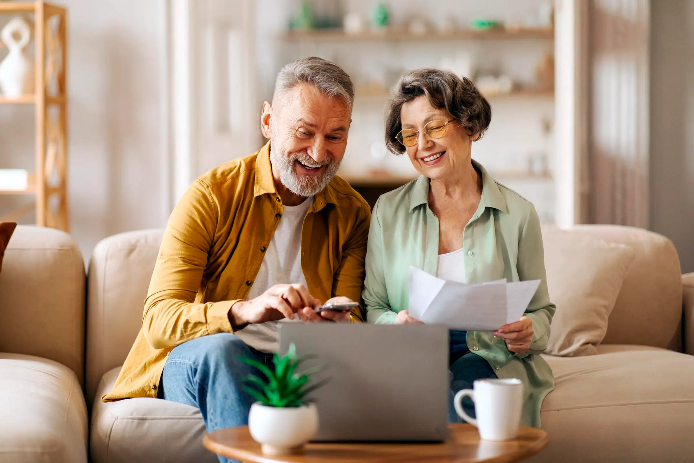 Older couple sitting on a beige couch in a bright living room, smiling and looking at a laptop on a wooden coffee table, the man holding a phone and the woman holding papers.