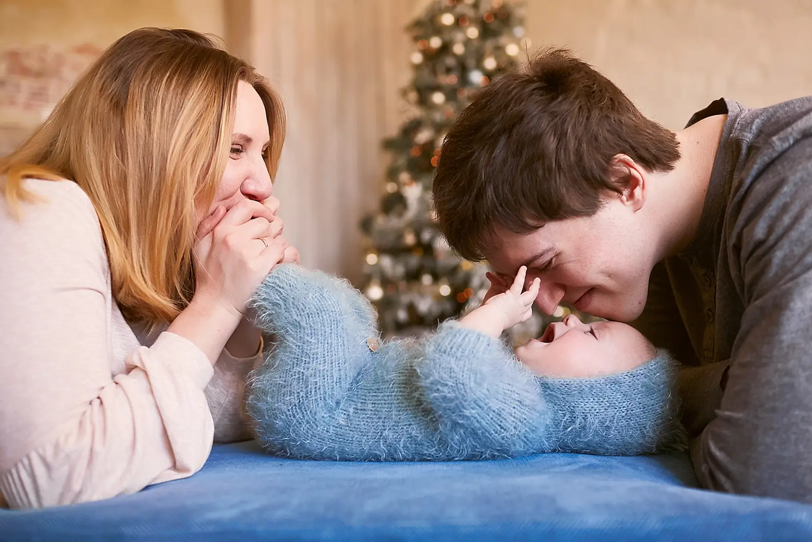 Smiling mother and father play with their baby lying on its back on a blue surface, with a decorated Christmas tree in the background.