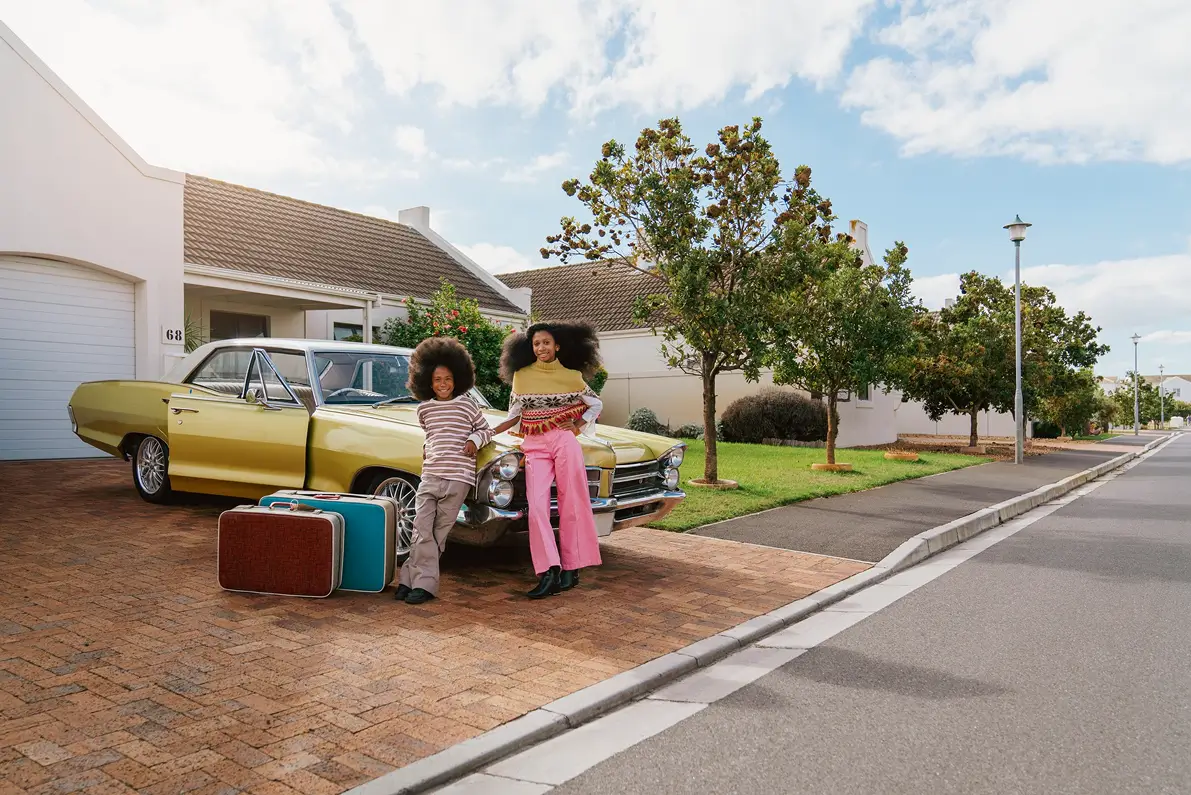 Two children with large curly hair leaning against the front of a yellow vintage car parked in a driveway, with two suitcases placed on the ground in front of the car, and suburban houses, trees, and a clear sky in the background.