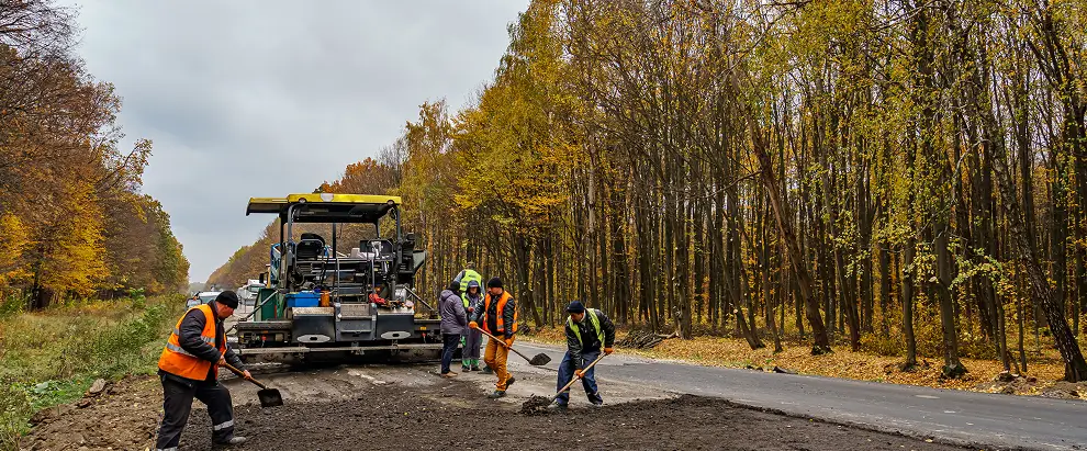 Construction workers in orange safety vests using shovels to spread asphalt on a road surrounded by autumn trees, with a large paving machine nearby under an overcast sky.