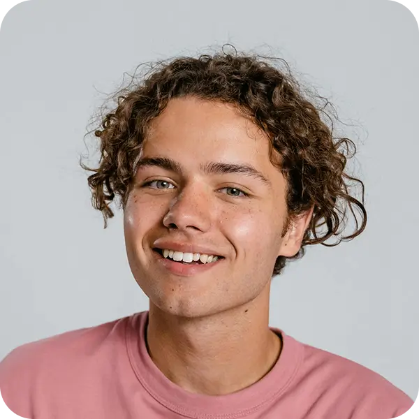 Young person with curly brown hair smiling, wearing a pink shirt against a plain light gray background.