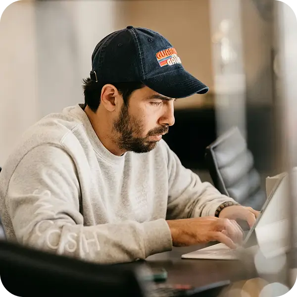 Bearded man in a gray sweatshirt and dark baseball cap focused on typing on a laptop in a modern indoor setting.