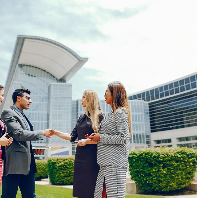 Four professionally dressed people engaged in a business handshake and conversation outside a modern glass office building.