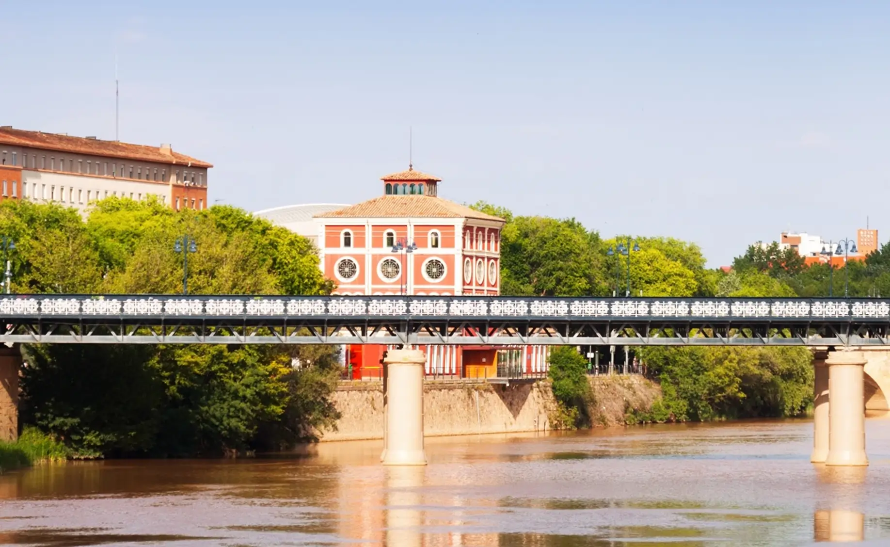 A metal bridge with decorative white railings spans over a calm river, with lush green trees along the riverbank and a distinctive red and white building with circular windows in the background.