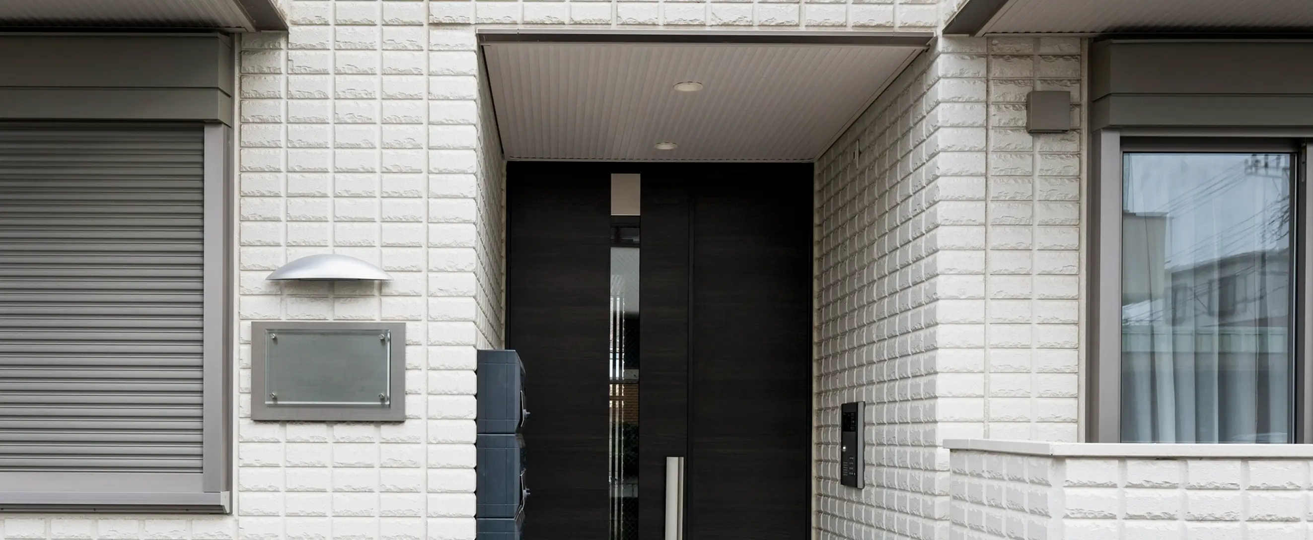 Modern house entrance with white brick walls, a dark double door with vertical glass panels, closed gray window shutters on the left, a window with curtains on the right, and an outdoor light fixture above a mailbox area.