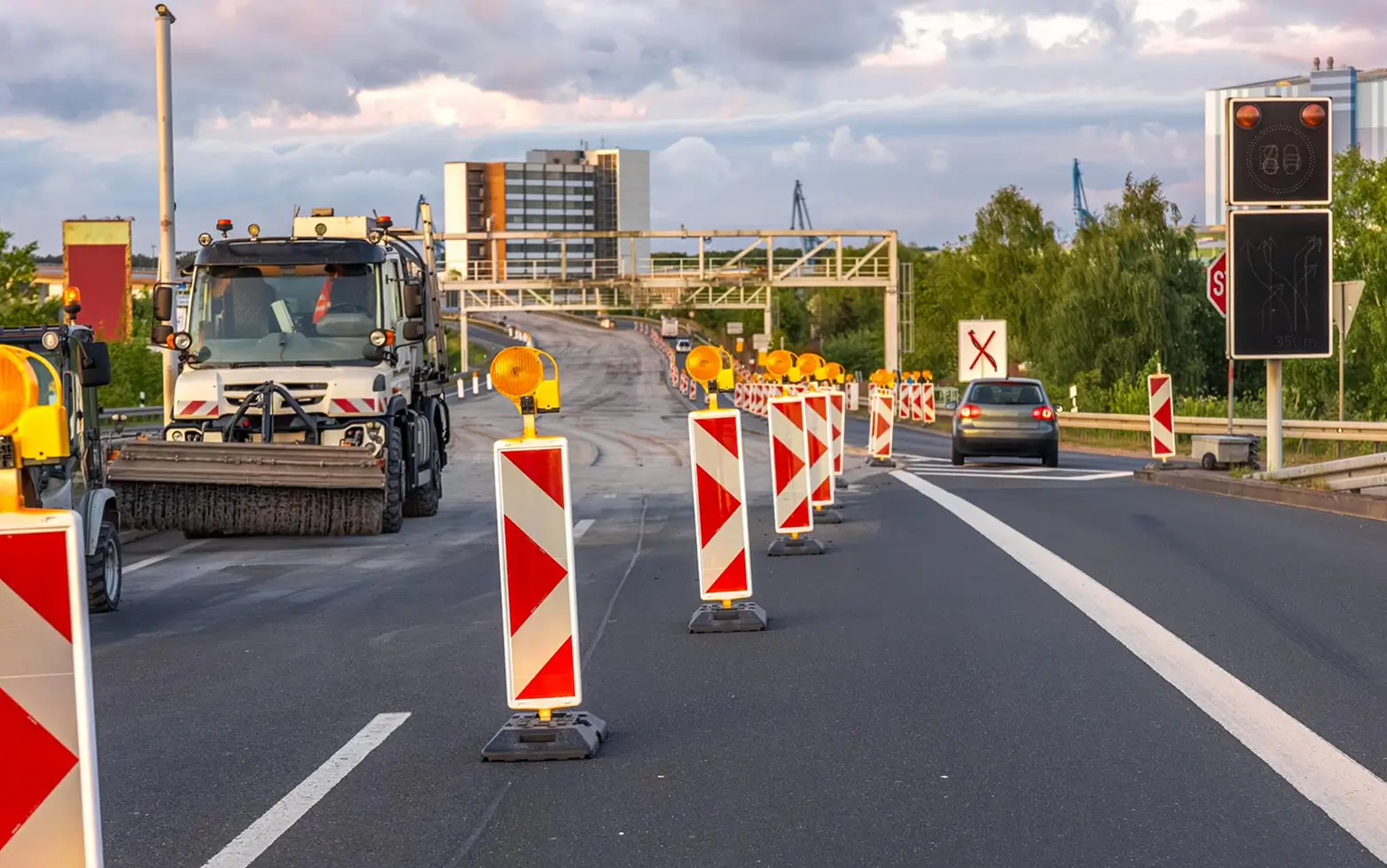 Street maintenance scene on a divided highway with a traffic barrier guiding cars to the right lane, roadwork equipment including a street sweeper, and multiple warning markers with orange lights during daytime under cloudy sky.