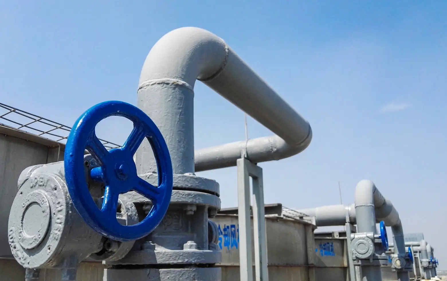 Close-up view of large industrial gray pipes with blue valve handles under a clear blue sky.