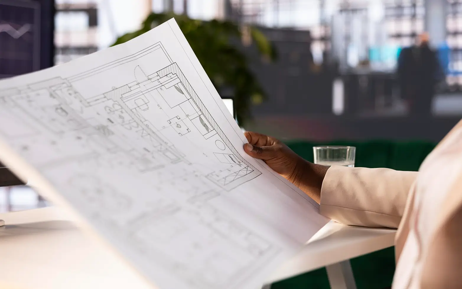 Close-up of a person holding and examining a large architectural blueprint or floor plan on white paper, with a glass of water on the desk in the background.