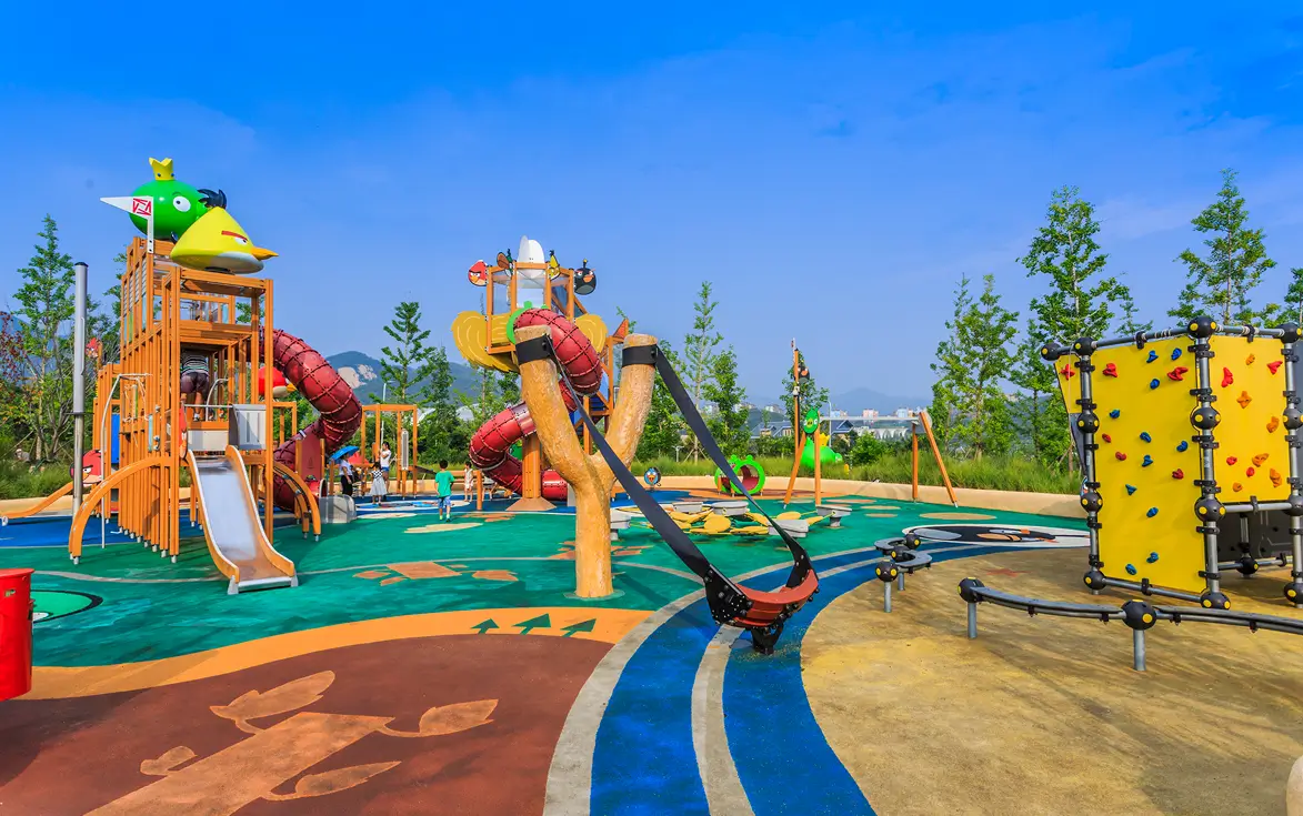 Colorful children's playground featuring a large wooden climbing structure with slides decorated like characters, a giant slingshot, climbing walls, and various play equipment on a vibrant safety surface under a clear blue sky.