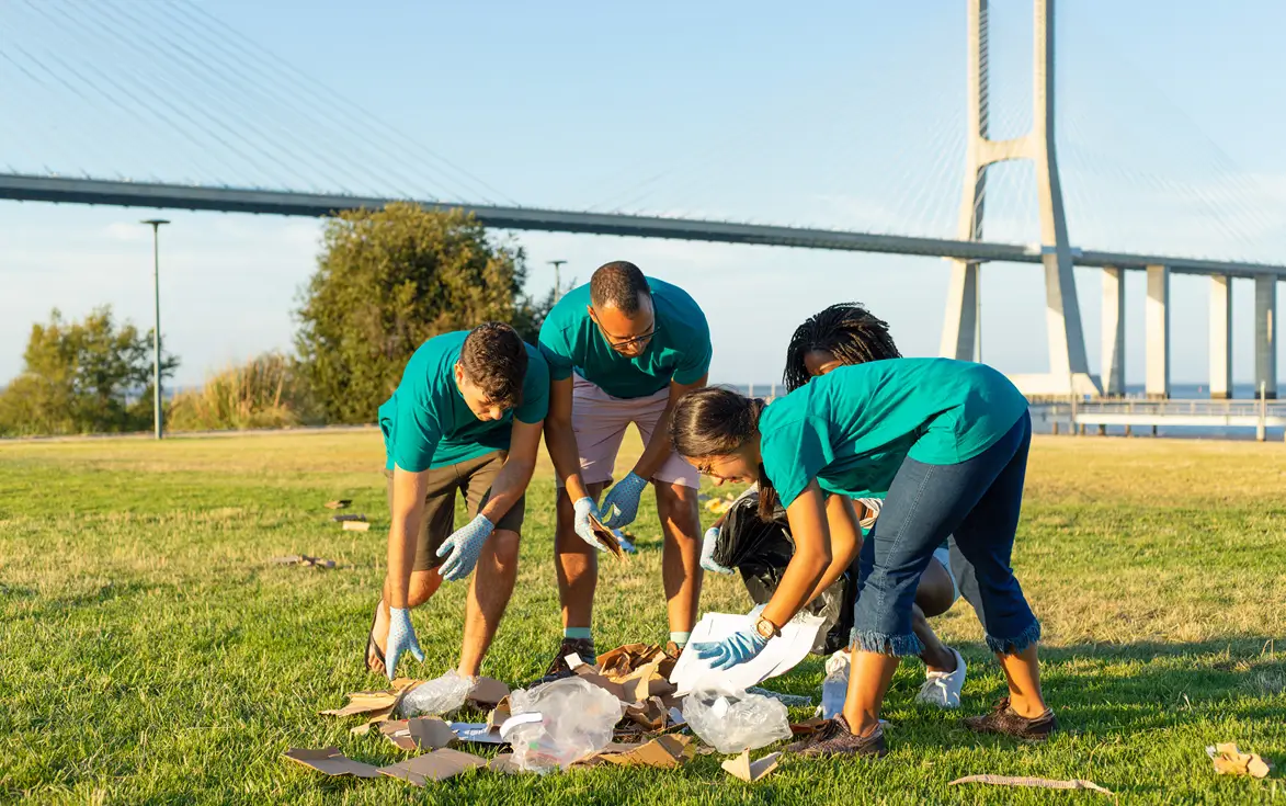 Four people wearing teal shirts and gloves are picking up litter on a grassy area near a large bridge during daylight.