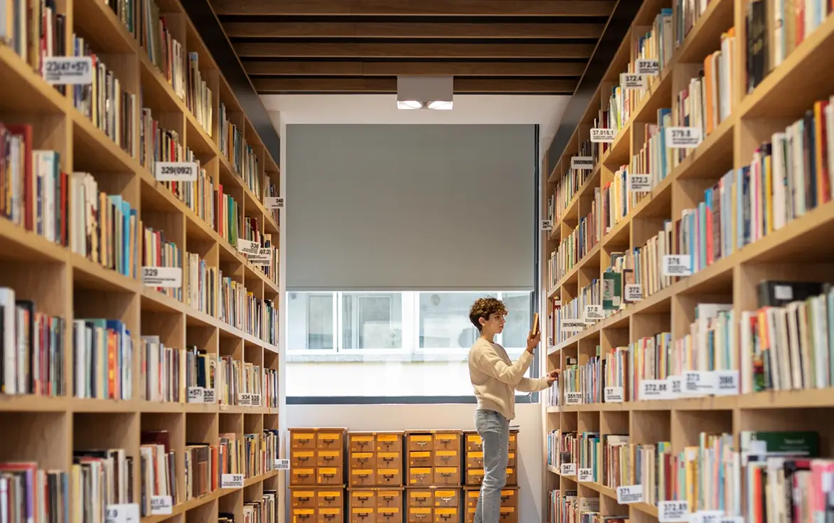 Person standing in a library aisle between tall wooden bookshelves filled with books, holding a book and looking at it, with a window and small yellow drawer units in the background.
