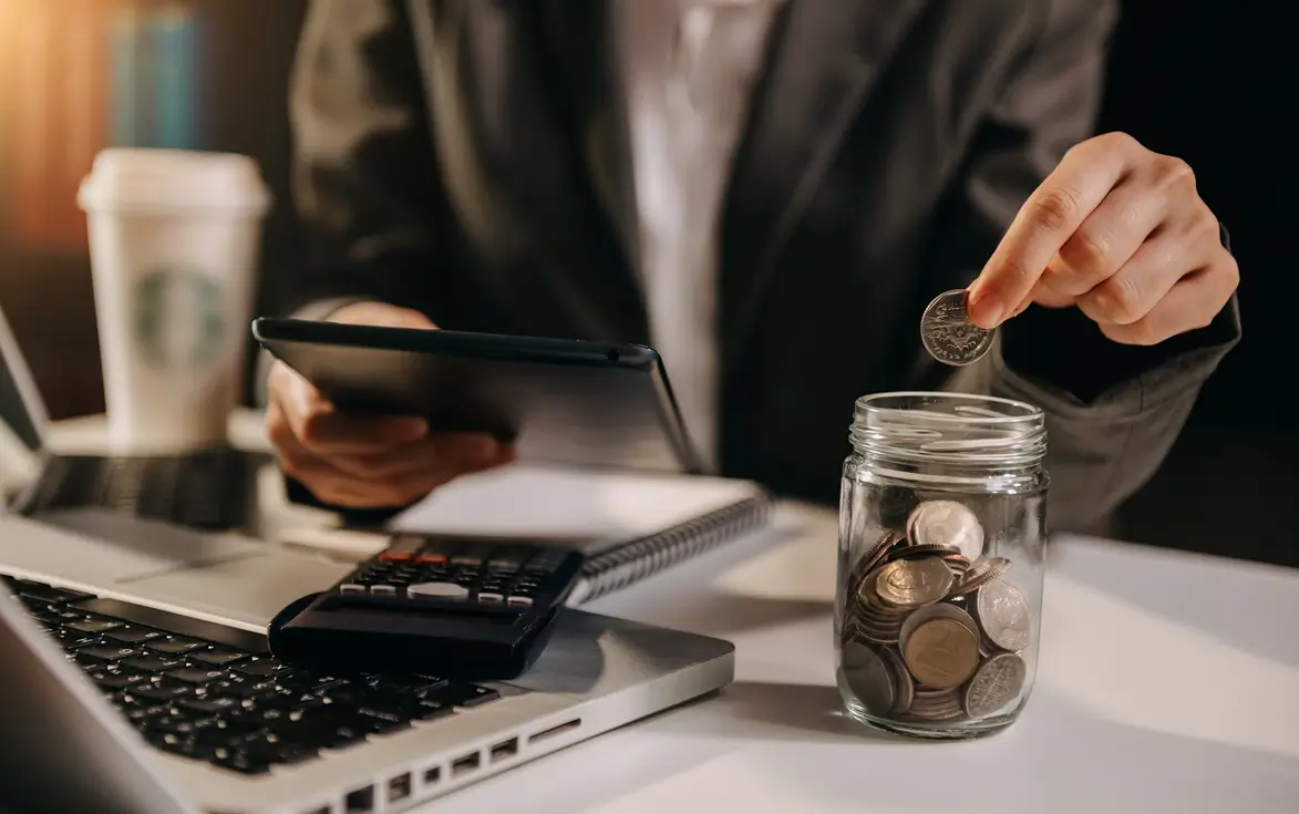 Person in a suit placing a coin into a glass jar filled with coins, sitting at a desk with a laptop, calculator, tablet, notebook, and a Starbucks coffee cup.