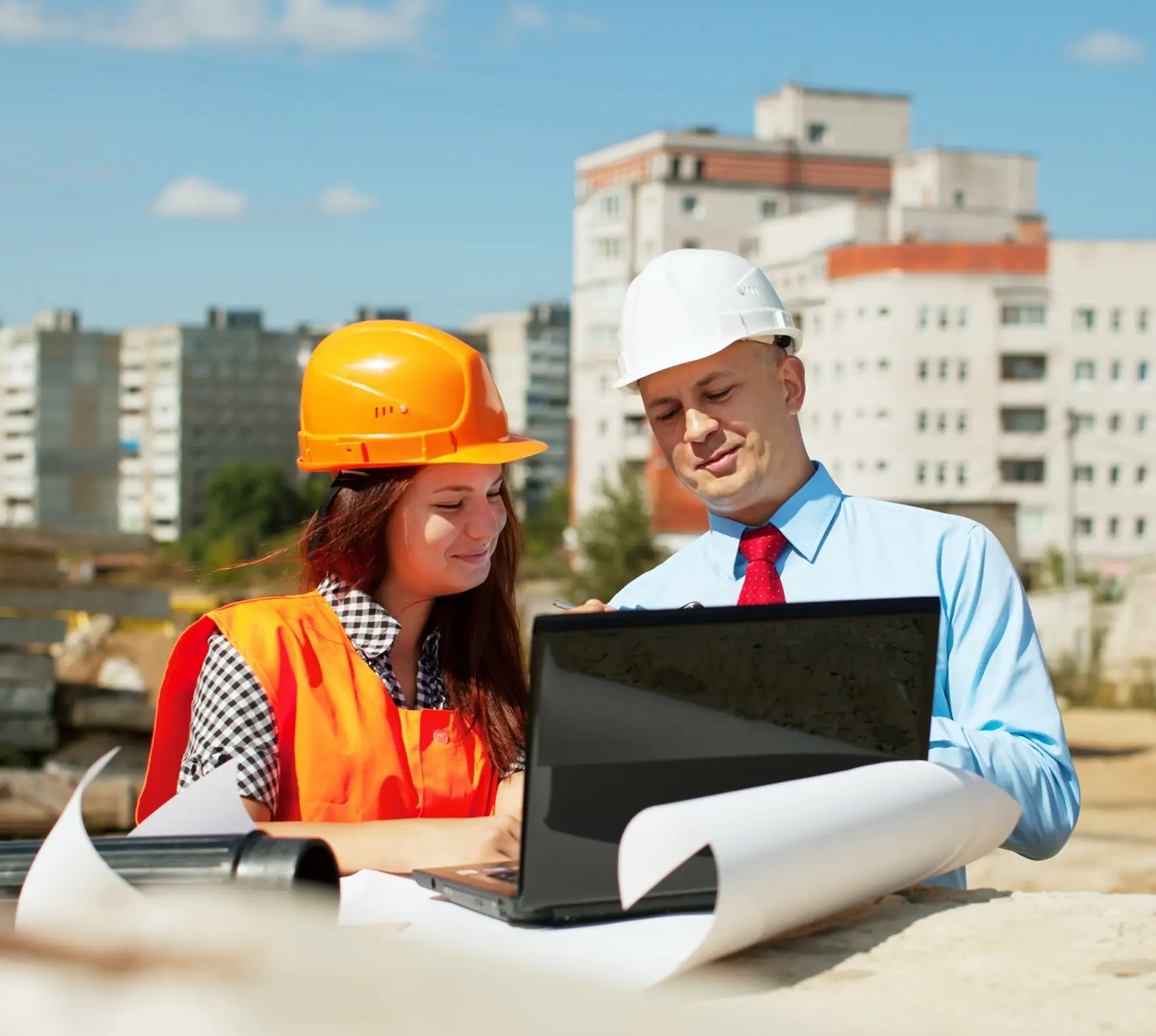 Two construction professionals, a woman in an orange safety vest and hard hat and a man in a white hard hat and blue shirt with red tie, looking at a laptop at a construction site with buildings in the background.