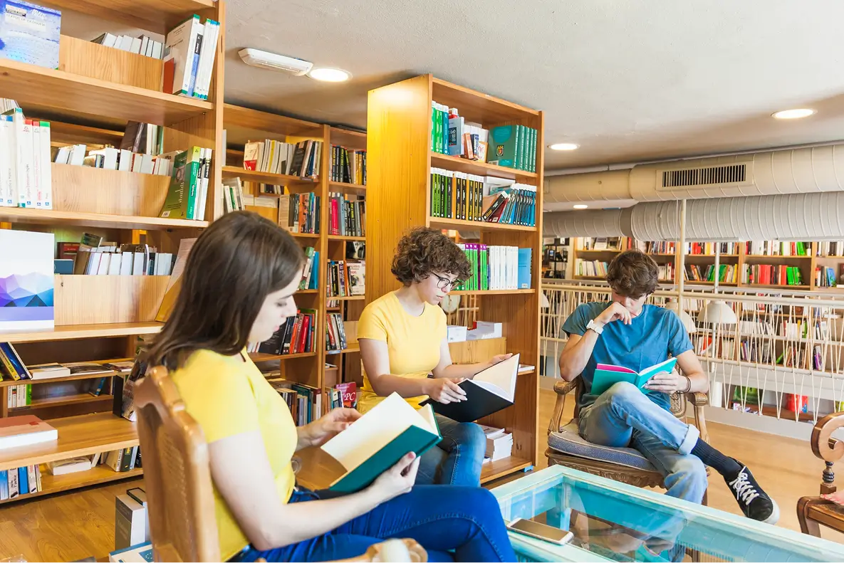 Three young adults seated in a cozy library room, reading books with wooden bookshelves filled with colorful books in the background, a glass table in front of them.