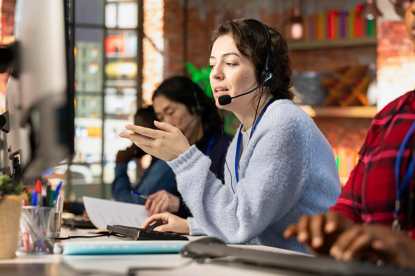 A woman wearing a headset speaking at a computer workstation in a busy office with other headset-wearing colleagues working in the background.