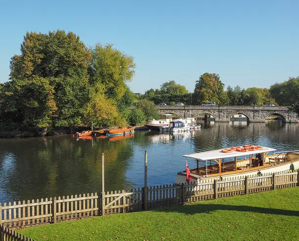 River scene with several small boats docked by a leafy riverside, a stone arch bridge in the background, and a grassy area with a wooden fence in the foreground under a clear blue sky.
