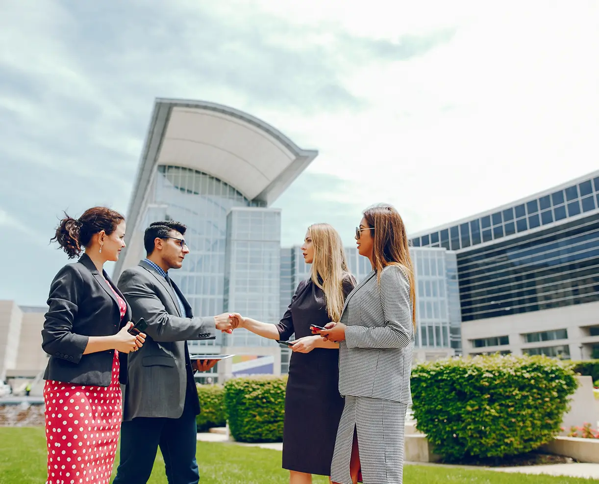 Two women and a man standing outside a modern office building with glass facades; the man is shaking hands with one woman while the other woman watches.