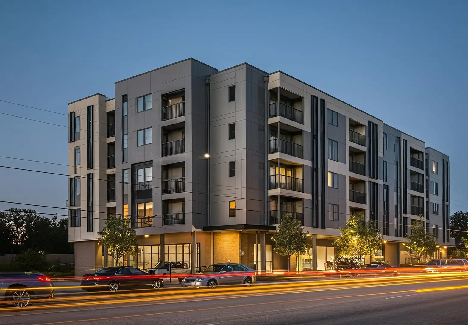 Modern multi-story apartment building at dusk with illuminated windows and balconies, street in front showing light trails from moving cars.
