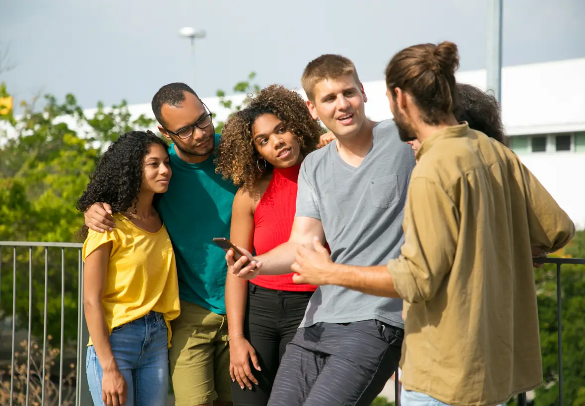 Group of five young adults standing outdoors, engaged in conversation; one man in a gray shirt holds a smartphone while another man in a beige shirt gestures as they talk, with three others closely listening and watching.