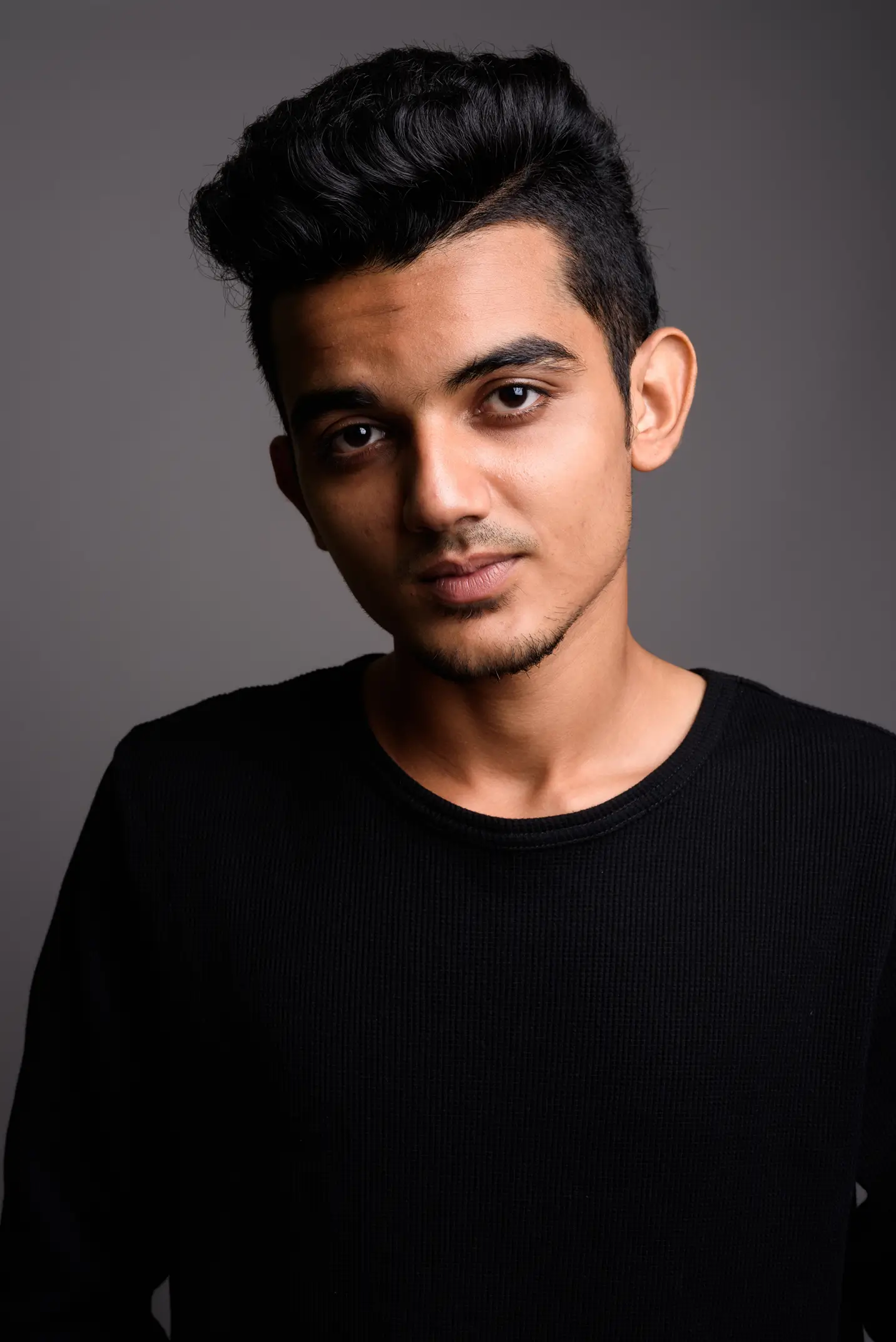 Portrait of a young man with dark hair and brown eyes wearing a black sweater against a plain gray background.