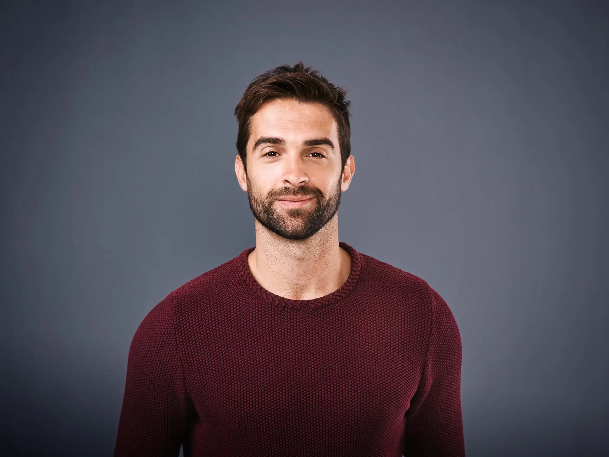 Smiling man with dark hair and beard wearing a maroon textured sweater against a plain gray background.