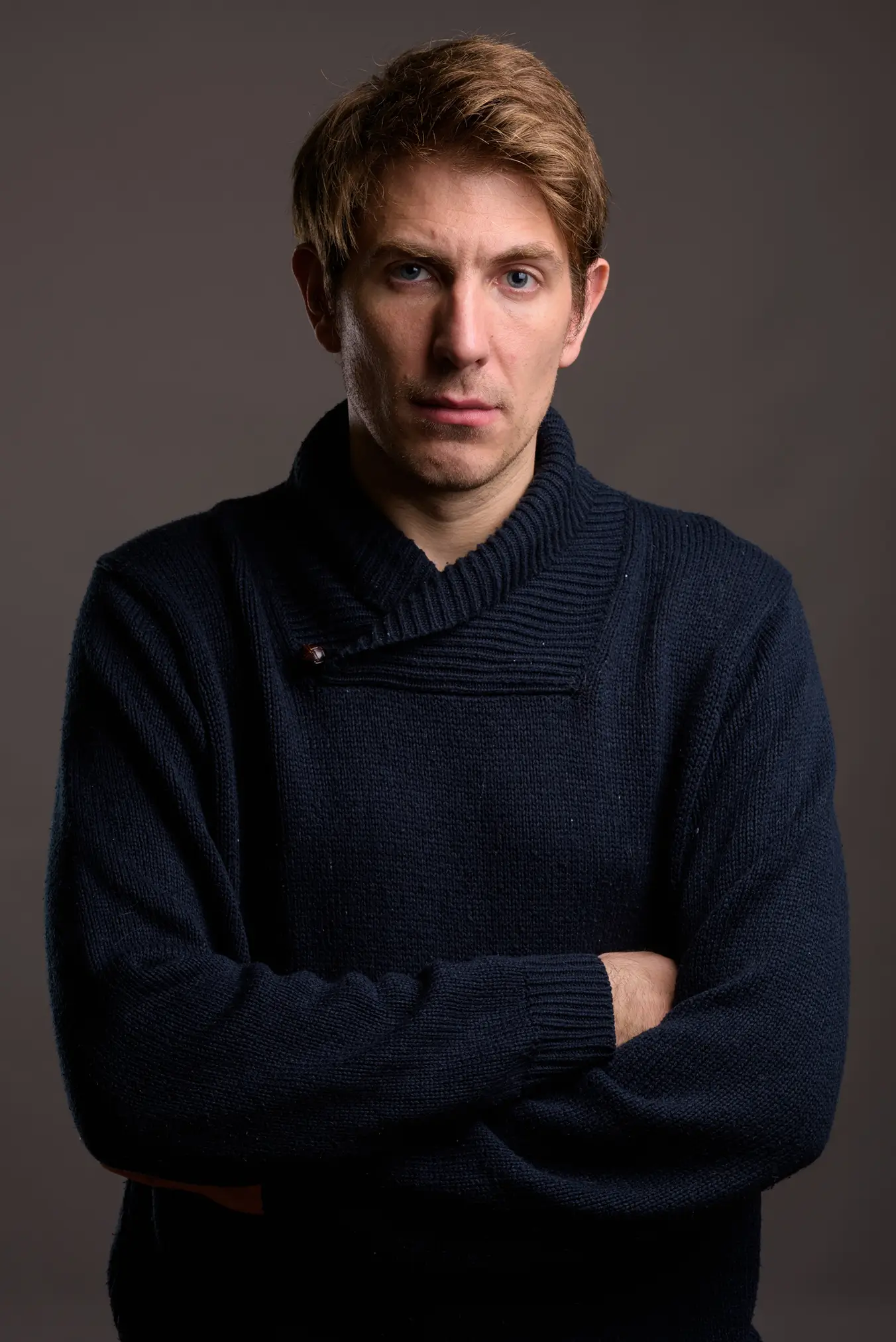 Portrait of a serious young man with light brown hair and blue eyes wearing a dark navy knit sweater with a high collar, arms crossed, against a plain dark background.