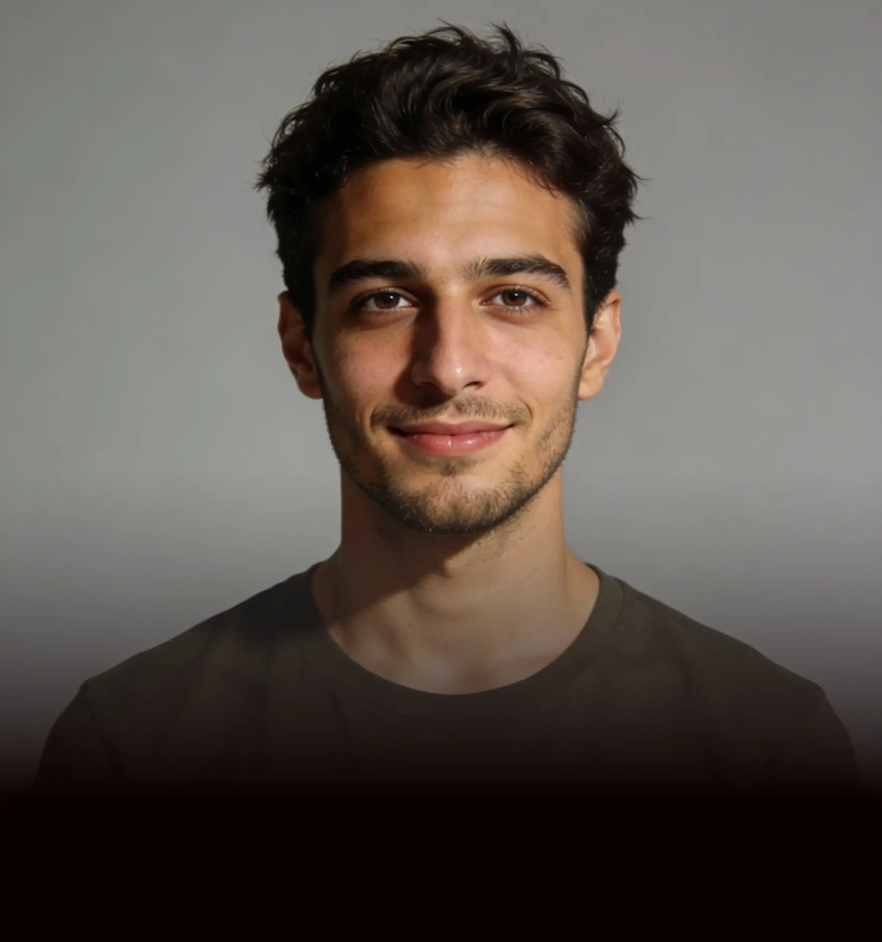 Portrait of a young man with short dark wavy hair, light facial hair, and brown eyes, wearing a dark olive-green shirt, smiling softly against a plain neutral gray background.