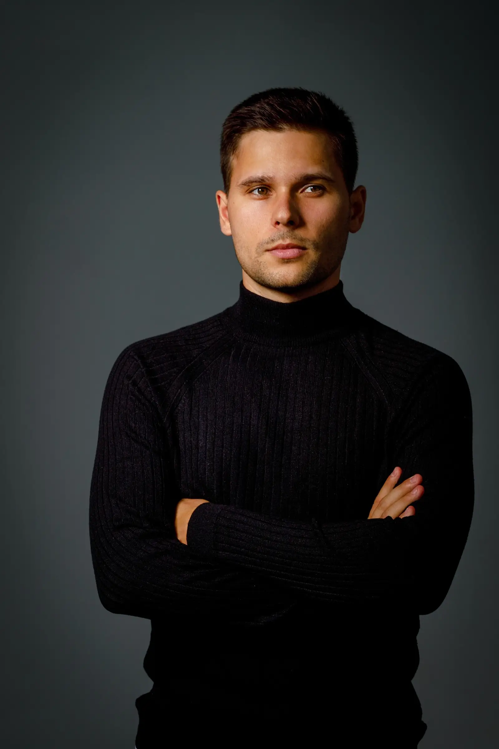 Portrait of a young man with short dark hair wearing a black ribbed turtleneck sweater, standing with arms crossed against a dark gray background, looking slightly to the side.