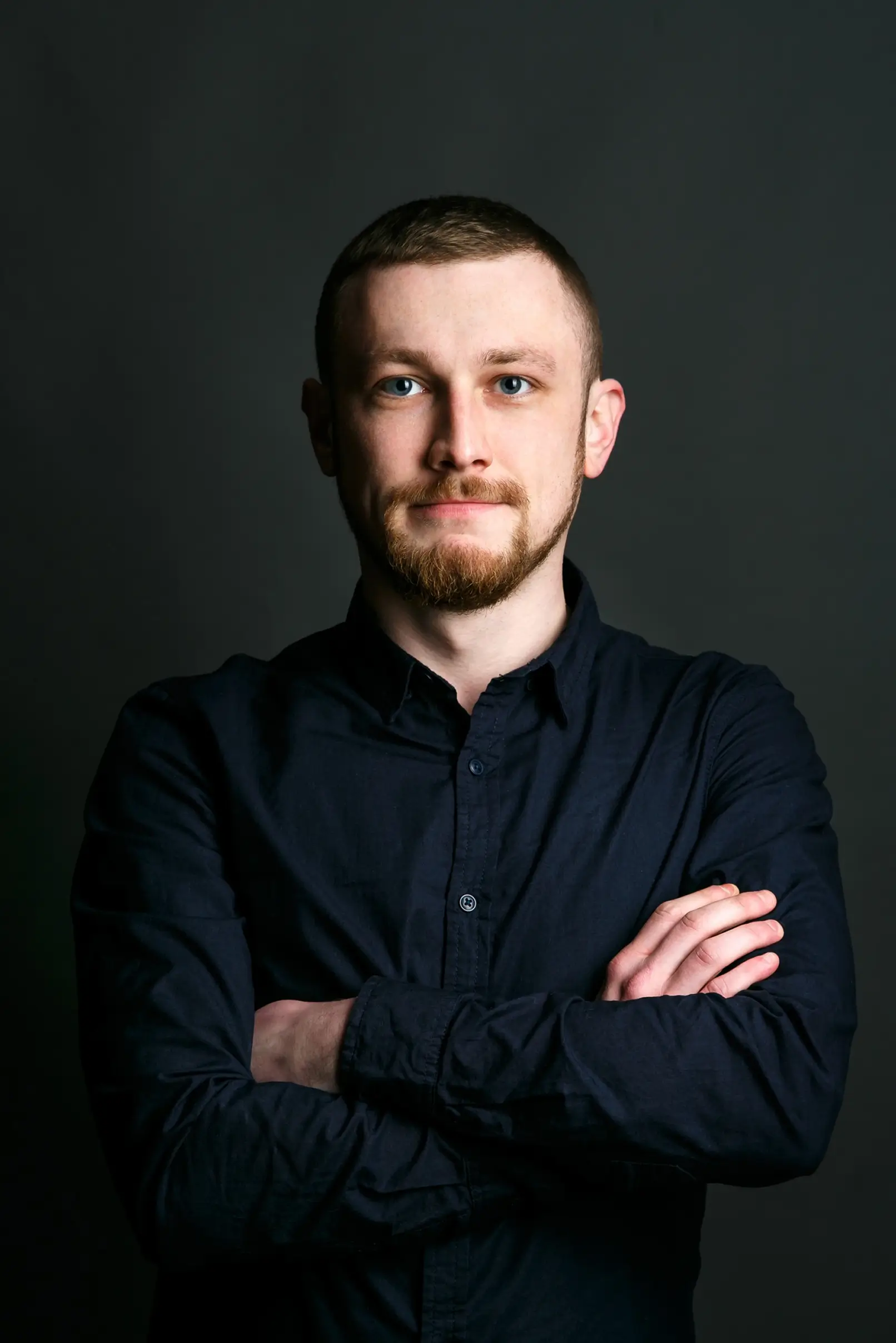Portrait of a young man with short brown hair and a beard wearing a dark button-up shirt, standing with arms crossed against a dark background.
