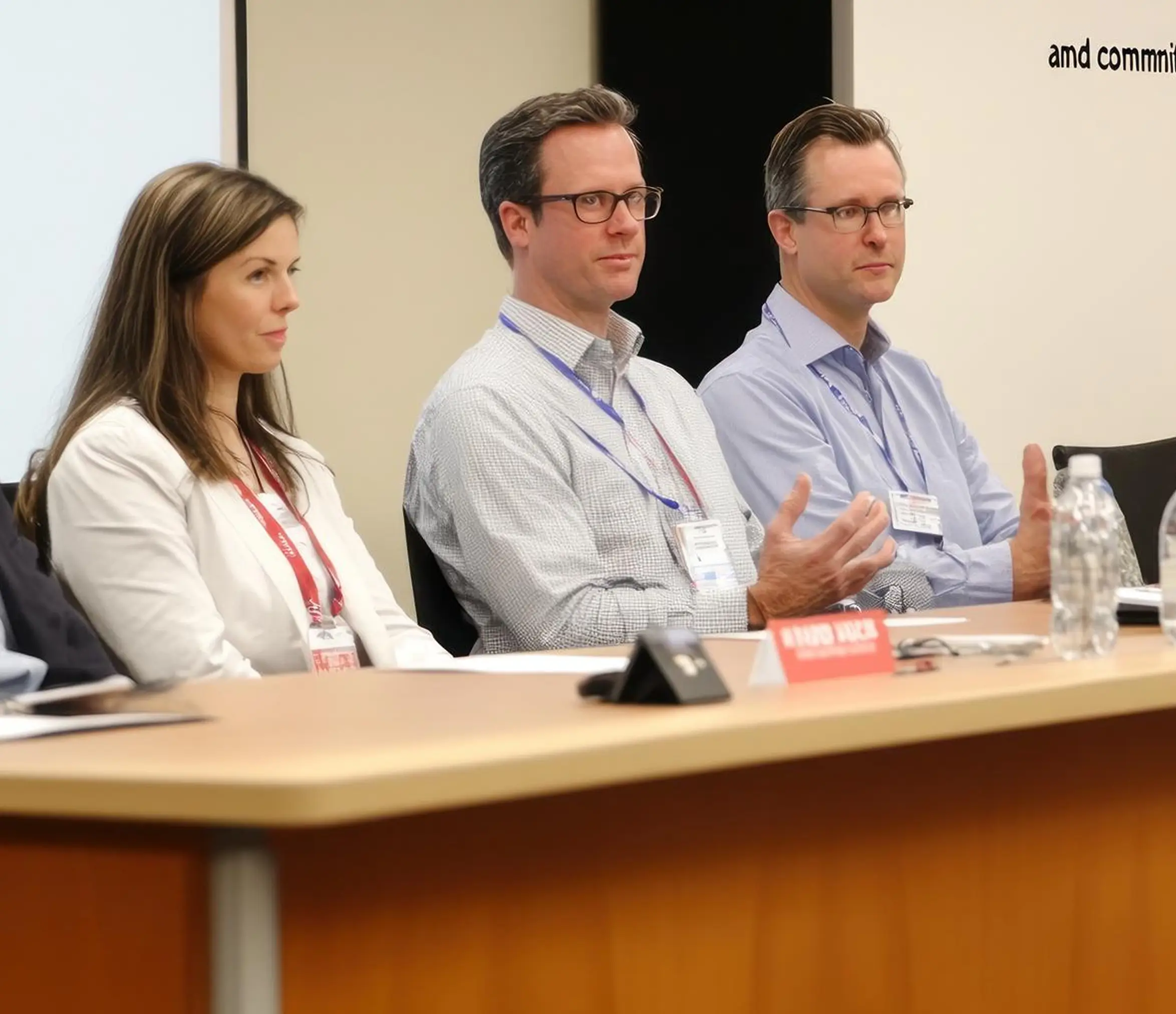 Three people sitting at a conference table during a meeting or panel discussion; a woman on the left wears a white jacket and a red lanyard, a man in the middle wears glasses with a white checkered shirt and blue lanyard, and another man on the right wears glasses with a light blue shirt and blue lanyard, gesturing with his hands.
