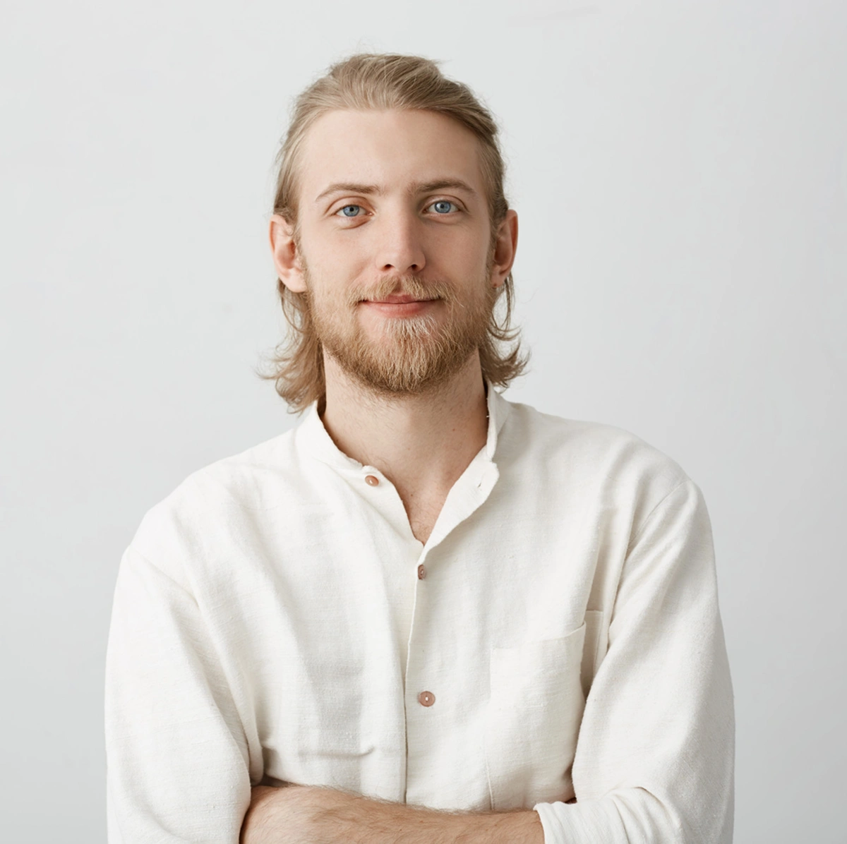 Smiling young man with light brown hair and beard, wearing a cream-colored button-up shirt, against a plain light gray background.