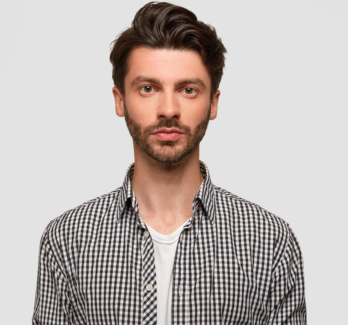 Man with light skin, brown hair, and beard wearing a black and white checkered shirt over a white T-shirt, looking directly at the camera with a neutral expression against a light gray background.