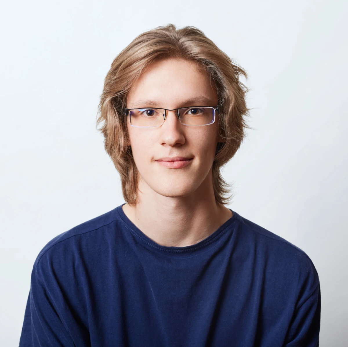 Young person with light brown hair and glasses, wearing a dark blue shirt, looking straight at camera with a neutral expression against a plain light background.