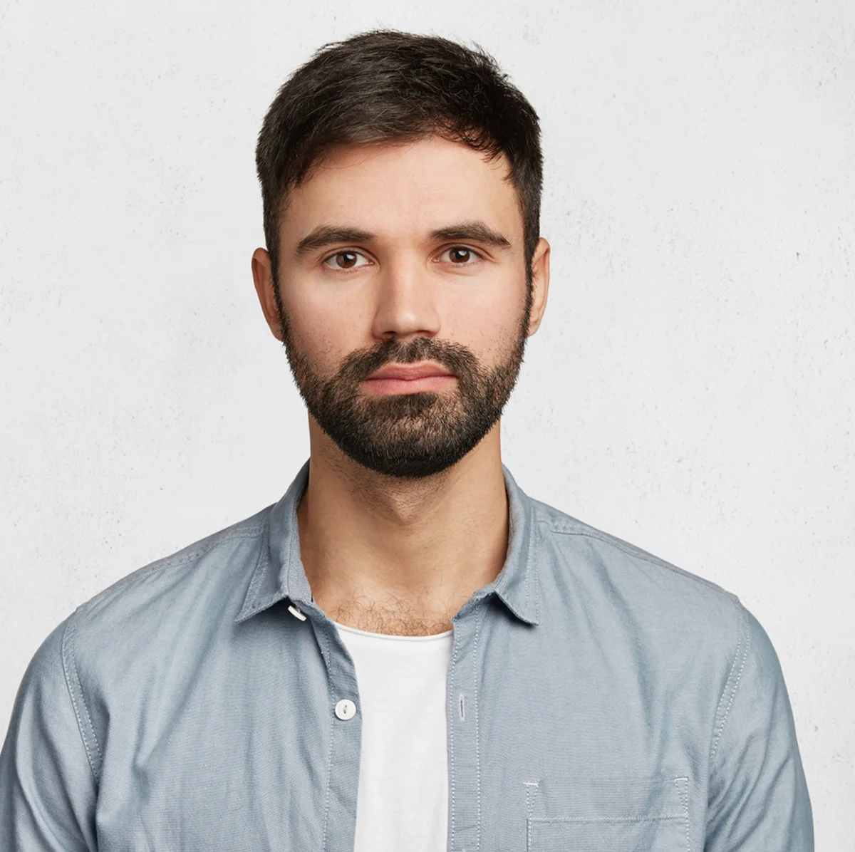 Portrait of a young man with short dark hair and beard, wearing a light blue button-up shirt over a white t-shirt, against a light grey background.