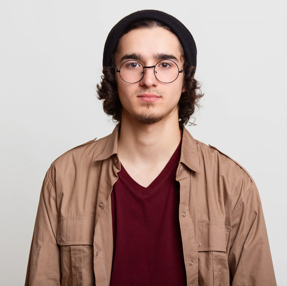Young man with curly hair wearing a black beanie and round glasses, dressed in a maroon v-neck shirt and a tan button-up jacket, looking straight ahead with a neutral expression.