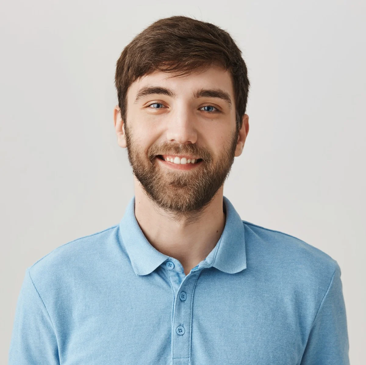 Smiling young man with short brown hair, blue eyes, and a beard, wearing a light blue polo shirt, against a plain light background.