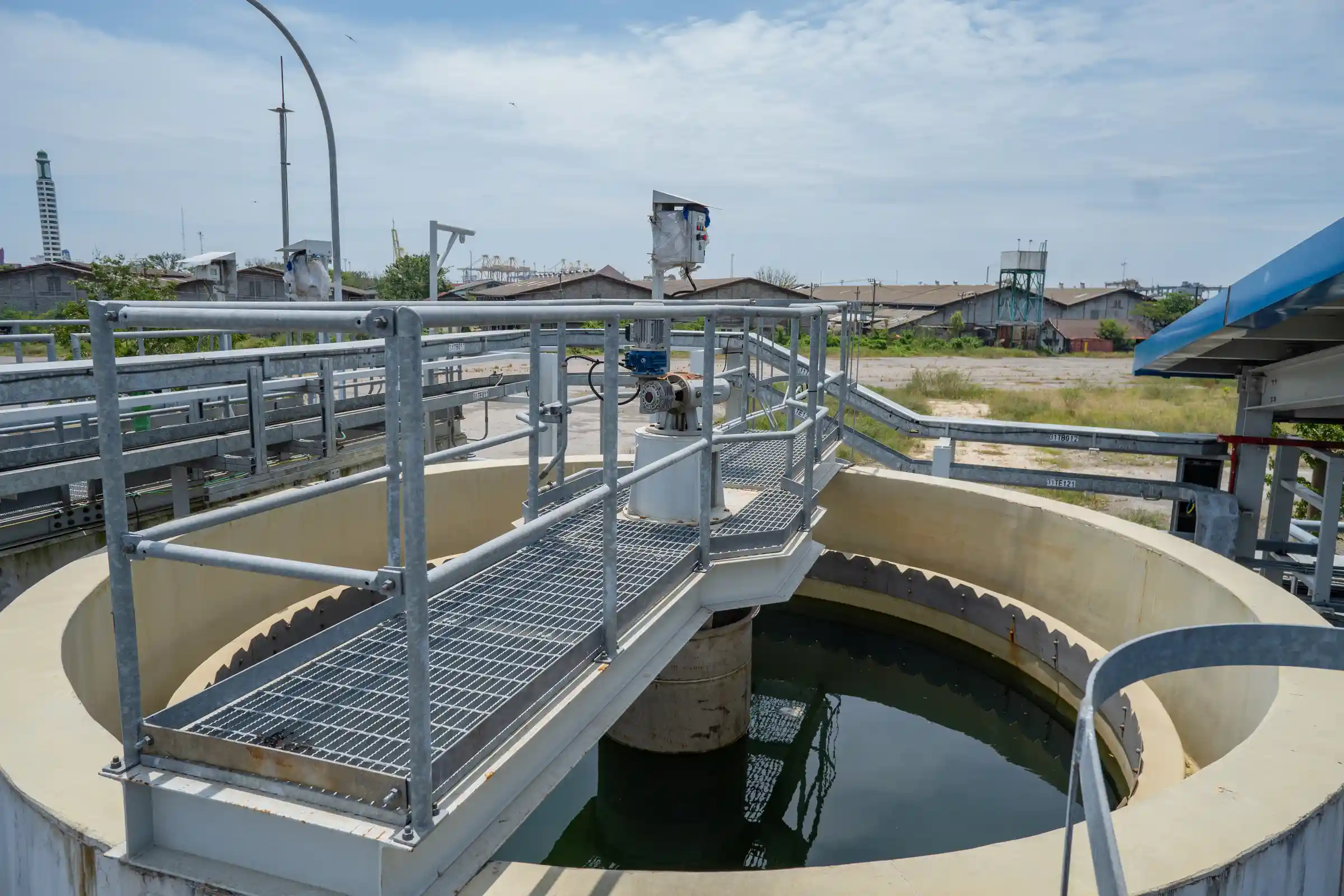 Circular water treatment tank with metal railing and walkway, outdoors under a cloudy sky, with industrial buildings and equipment in the background.