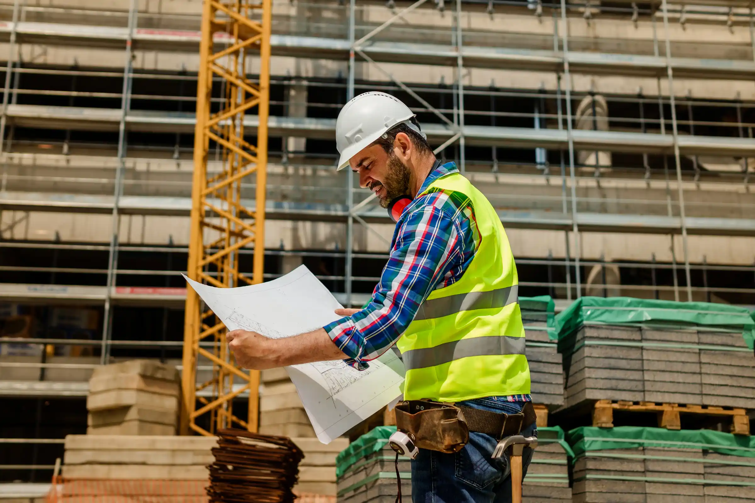 Construction worker wearing a white hard hat and yellow safety vest examines building plans at a construction site with stacked materials and scaffolding in the background.