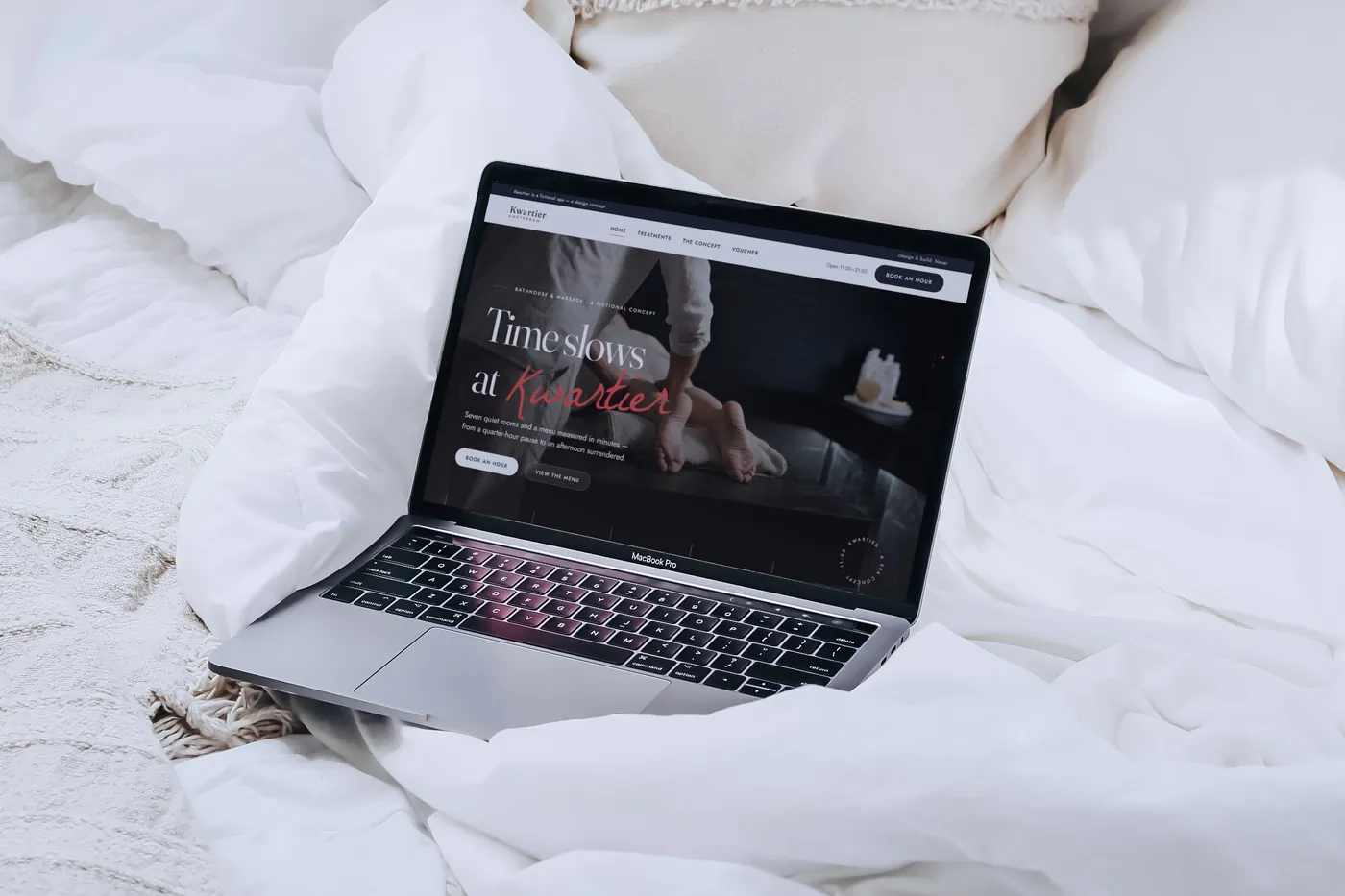 The Kwartier concept homepage on an open MacBook Pro among white sheets and a knitted throw on a bed.