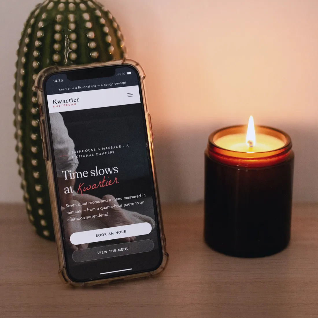 The Kwartier concept homepage on an iPhone in a clear case, leaning against a ceramic cactus beside a lit candle.