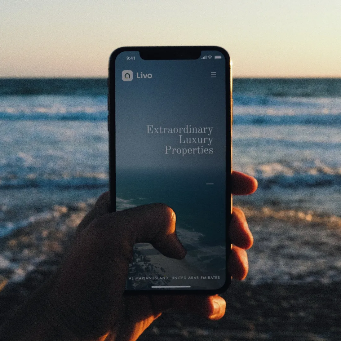 The Livo concept homepage on an iPhone held up against the sea at sunset.