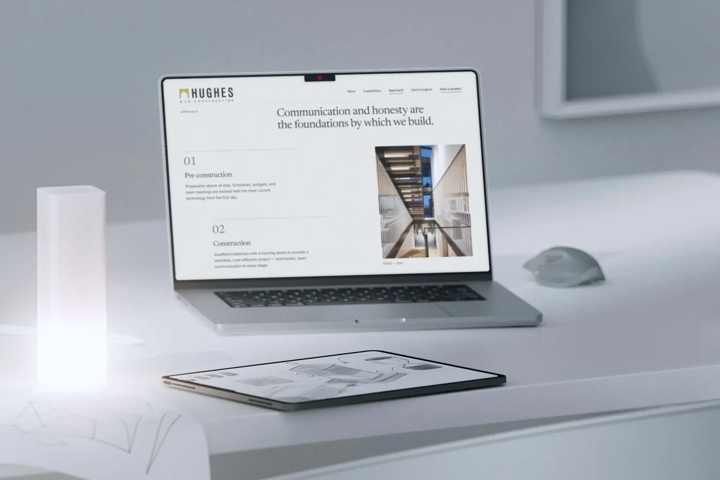 The approach section of our Hughes & Co. redesign concept on a MacBook Pro on a white desk, beside a glowing lamp and an iPad with architectural sketches.