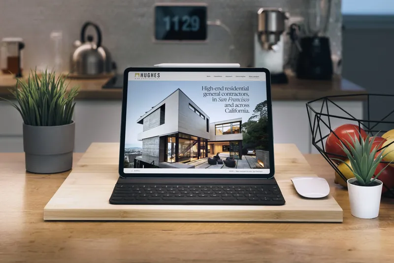 Our redesign concept for Hughes & Co. Construction on an iPad in a keyboard case, standing on a wooden board on a kitchen counter.