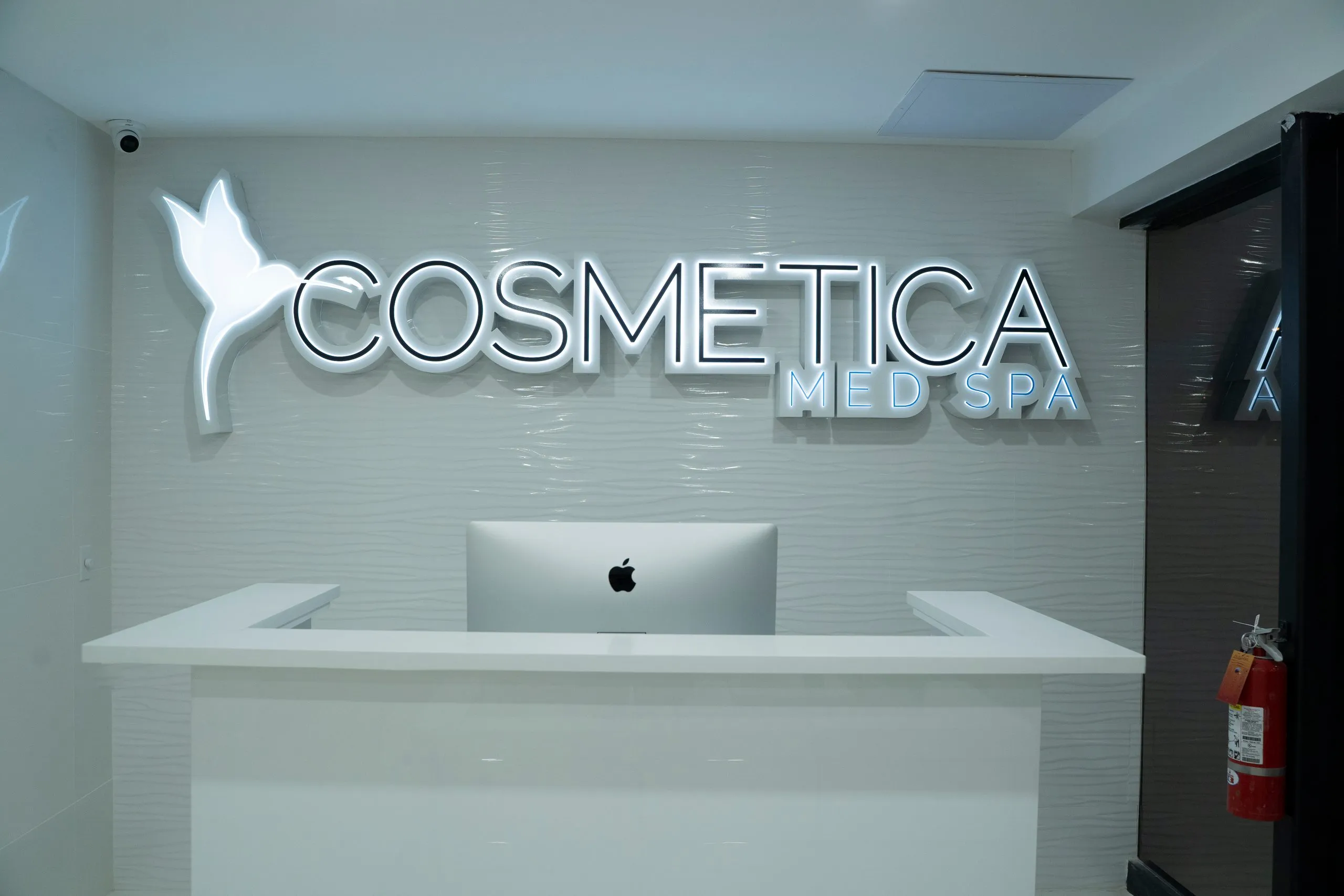 Reception counter and backlit logo wall beside the glass entrance at Cosmetica Med Spa
