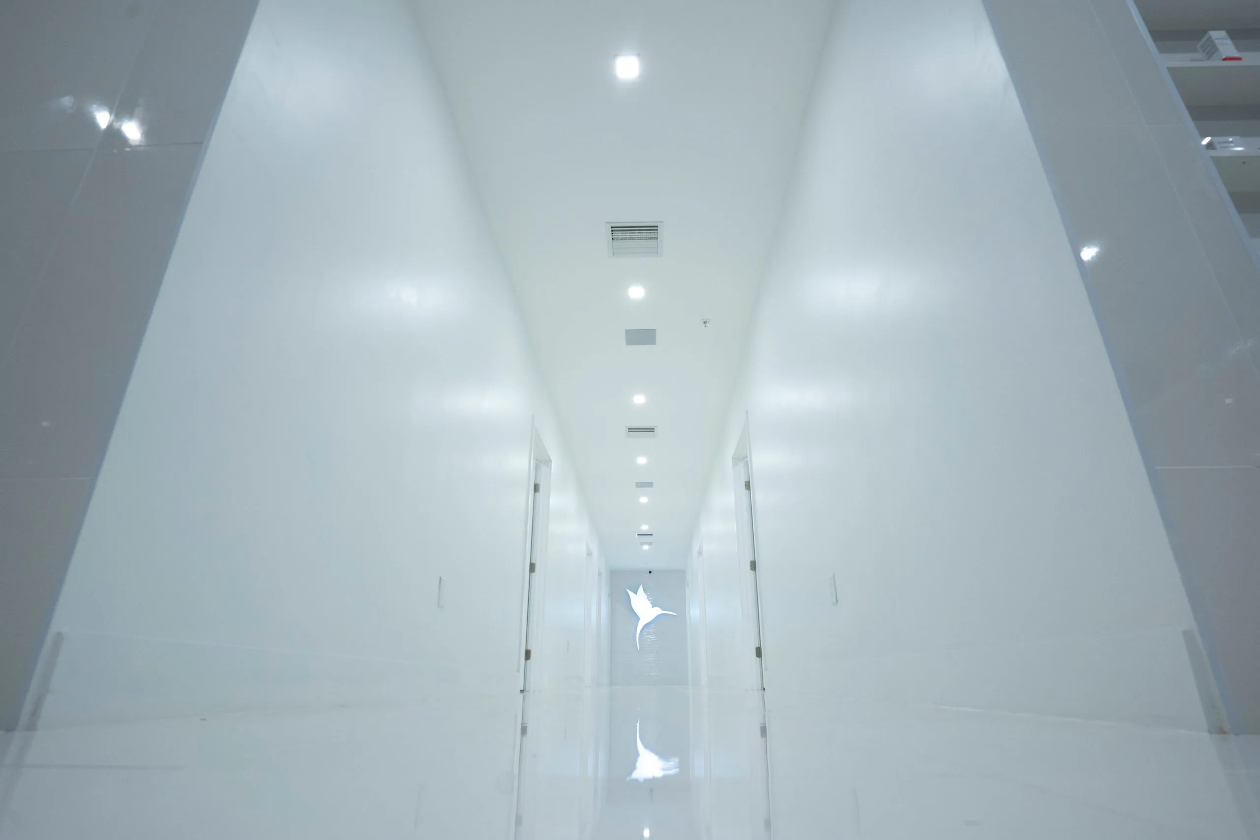 Low-angle view of a long white corridor ending in the glowing hummingbird logo at Cosmetica Med Spa