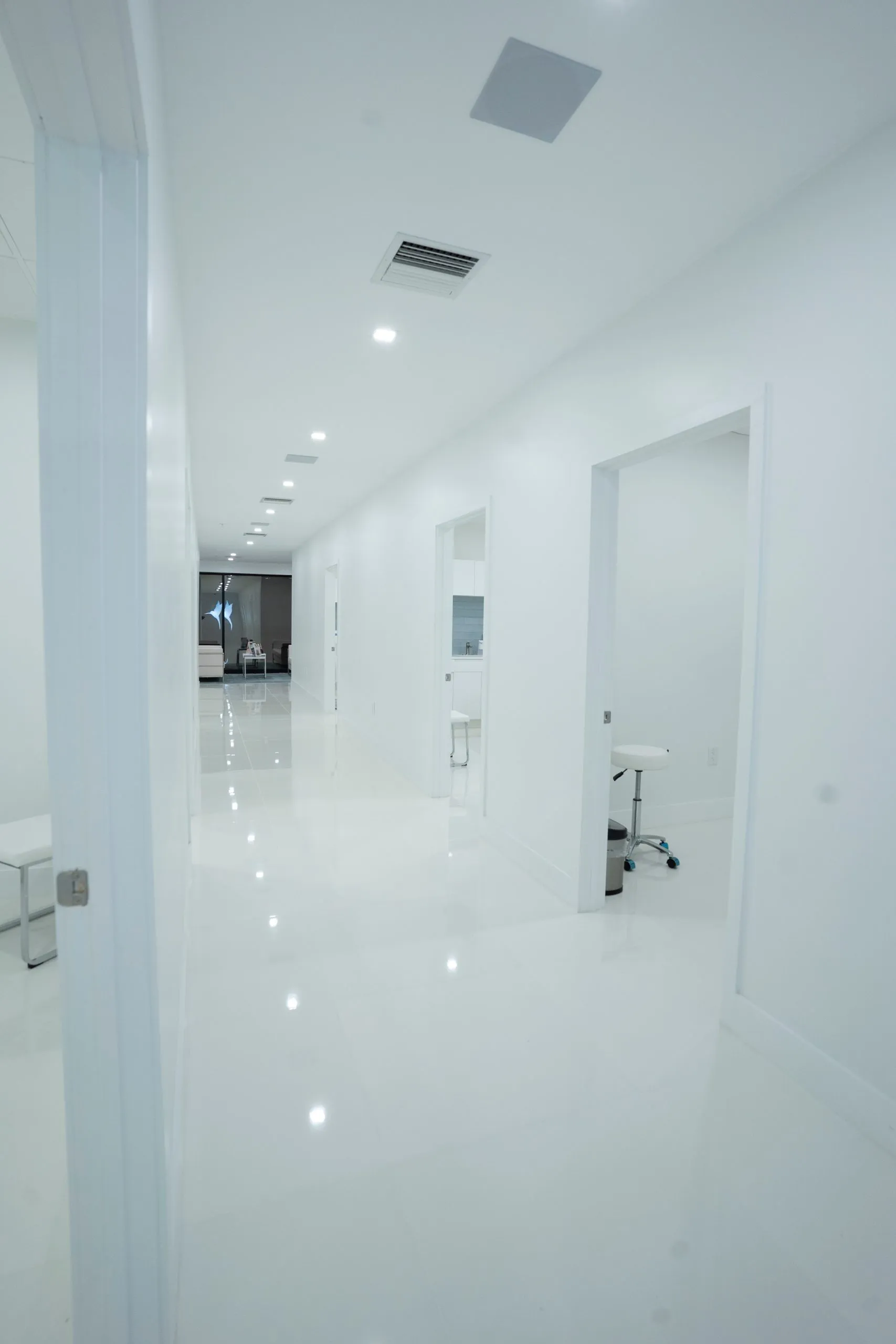 Glossy white hallway with open treatment room doors leading to the lobby at Cosmetica Med Spa